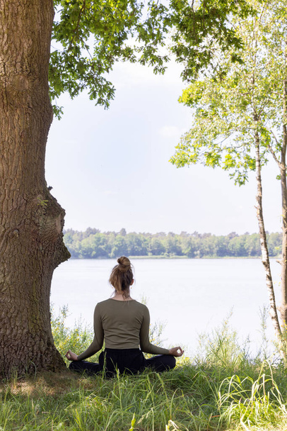 Junge Frau praktiziert Yoga in der Natur. Auf grünem Gras neben einem Baum und einem Waldsee sitzend. Kommunizieren Sie, um sich zu bewegen und gesund zu bleiben. Hochwertiges Foto - Foto, Bild