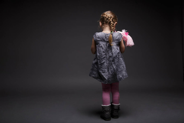 Back view of a little girl with doll, toddler, offended by someone, being in bad mood, posing isolated over dark grey background. - Foto, immagini