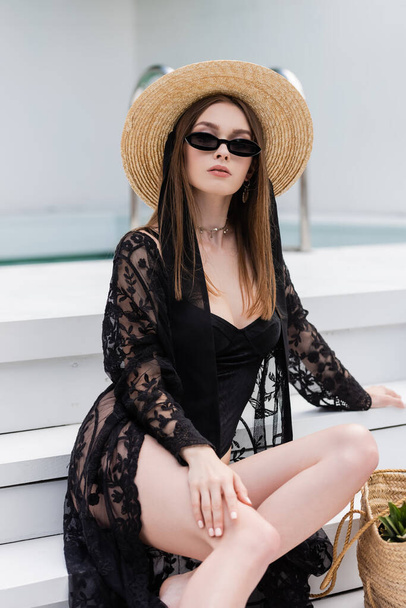 Stylish young woman in swimsuit and sunglasses sitting near handbag on stairs at resort  - Photo, image