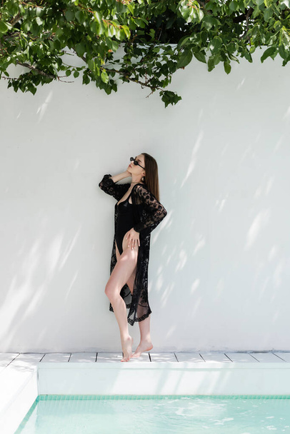 Stylish woman in guipure robe and swimsuit standing near wall and swimming pool  - Foto, afbeelding