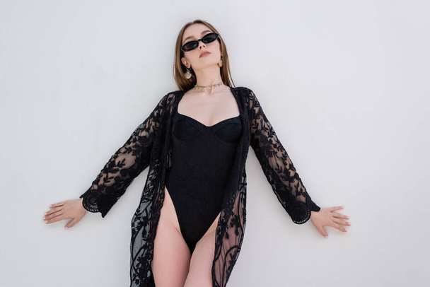 Trendy woman in black robe and swimsuit posing near wall at resort  - Photo, image