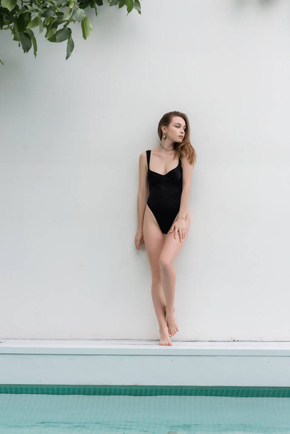 Stylish woman in swimsuit standing near wall and swimming pool at resort  - Valokuva, kuva