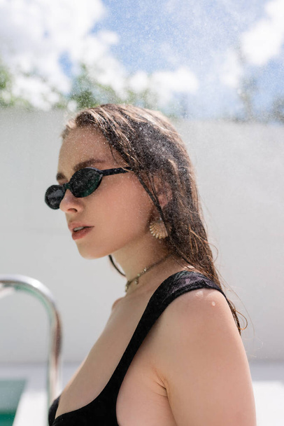 Trendy young woman in sunglasses standing under water drops at resort  - Fotó, kép