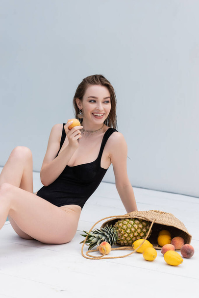 Cheerful young woman in swimsuit holding peach near fruits in straw handbag at resort  - Photo, Image