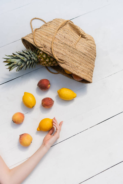 Top view of woman touching fresh lemon near fruits in straw handbag on floor  - Φωτογραφία, εικόνα