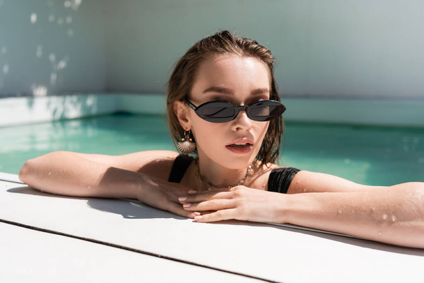 portrait of stylish woman in black sunglasses and golden accessories resting in swimming pool  - Fotó, kép