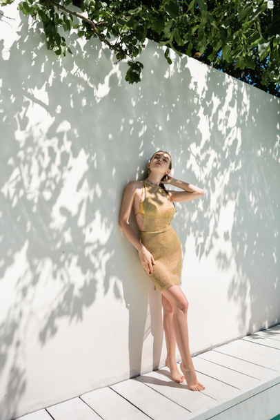 full length of barefoot young woman in golden summer dress standing near while wall with shadows from green leaves - Fotoğraf, Görsel