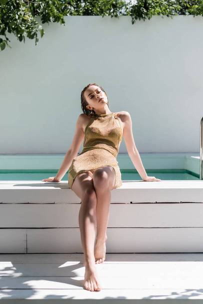 full length of barefoot and stylish woman in golden dress and swimwear sitting at poolside  - 写真・画像