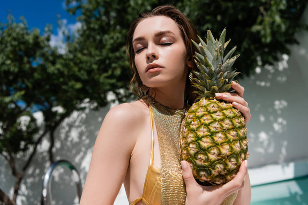 low angle view of young woman with closed eyes holding ripe pineapple outside - Photo, Image