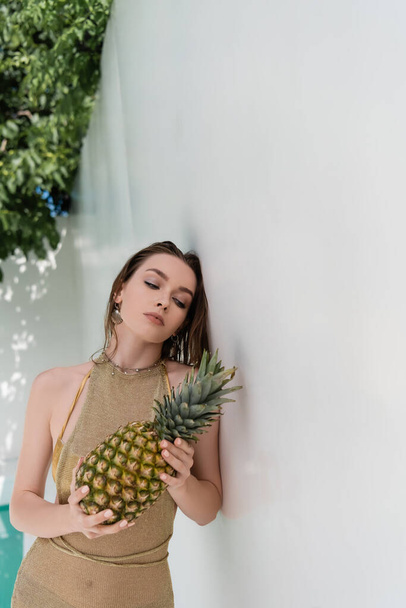 pretty young woman in golden summer dress holding ripe pineapple near white wall - Foto, Imagem