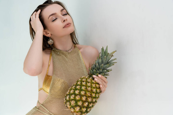 young woman in golden summer dress holding ripe pineapple near white wall - Fotó, kép
