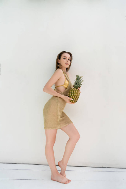 full length of young woman in golden summer dress holding fresh pineapple near white wall - Foto, Imagen
