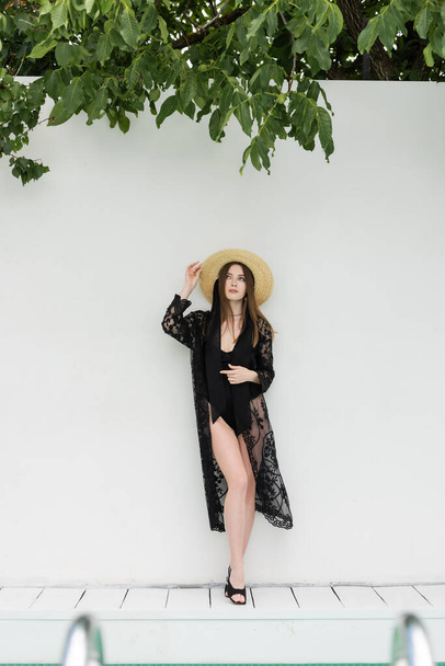 stylish young woman adjusting straw hat while standing in black knitted robe near white wall  - Fotoğraf, Görsel