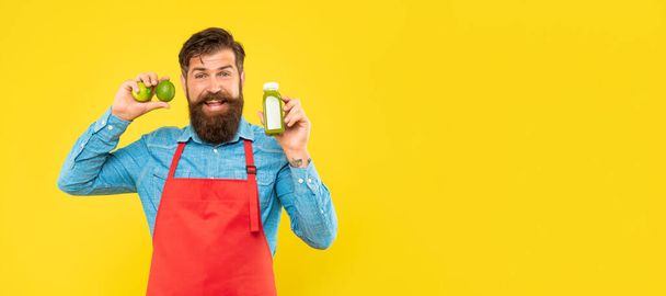 Happy man in apron holding limes and juice bottle yellow background copy space, juice barkeeper. - 写真・画像