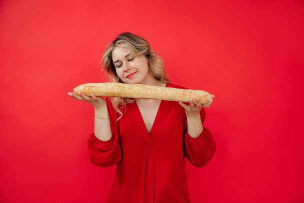 Smiling glad and calm attractive blonde woman in red dress holding gently and smelling french bread baguette in hands on red studio isolated background. Tasty calorie food on holidays. Copy space - Foto, Imagem