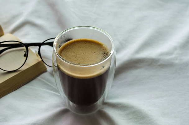 Een glas warme koffie met boek en bril op bed in de ochtend. Blijf thuis en ontspan je.. - Foto, afbeelding
