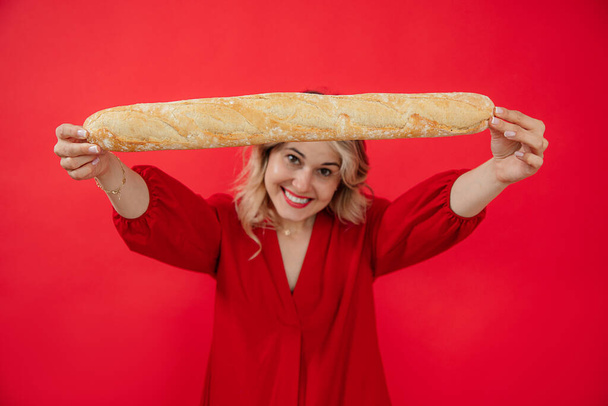 Smiling glad and joyful blonde woman in red dress holding and showing long french bread baguette in hands on red studio isolated background. Promotion of snacks, tasty, delicious food for the market - Foto, Bild