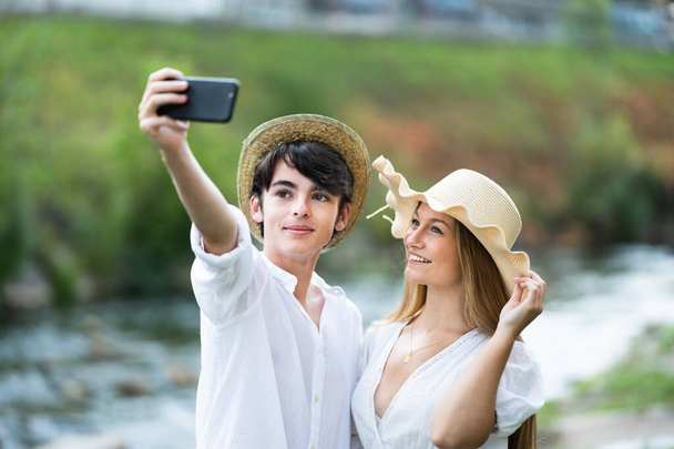 Happy teenager tourist couple looking at phone and taking selfie in nature, beside river. - Foto, imagen