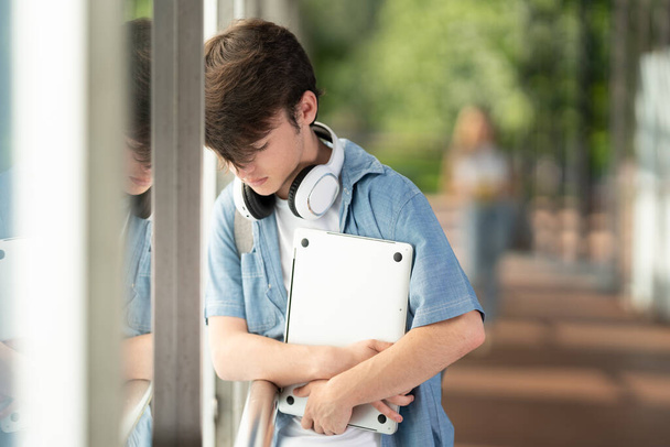 Rapaz adolescente triste segurando laptop contra parede em um corredor na faculdade. Conceito de depressão - Foto, Imagem