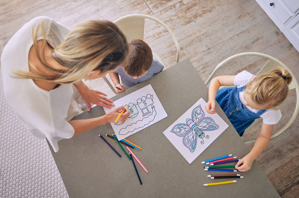Above shot of little girl and boy sitting at table with colourful pencils and pictures while colouring with mom helping. Caucasian mother with two kids enjoying educational pastime and being creative. - Foto, afbeelding