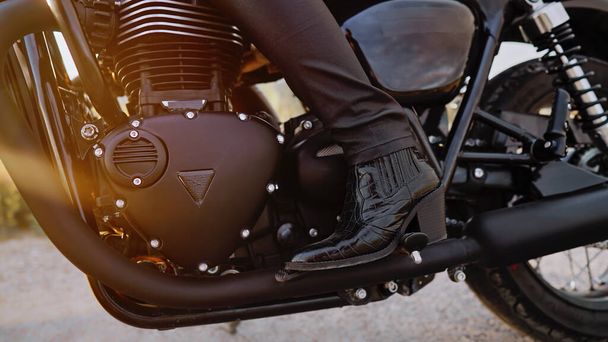 Legs of stylish motorcyclist woman sitting on classic bike. Black retro-styled motorcycle. Details of vintage design of brand-new motorbike. Sun flare, outdoor. High quality photo - Фото, изображение