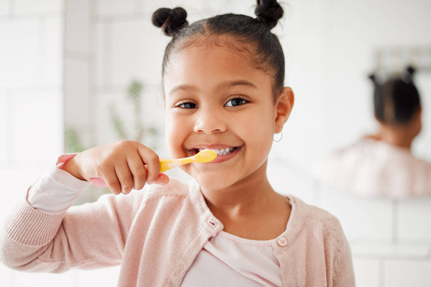 Μια μικτή φυλή αξιολάτρευτο κοριτσάκι βουρτσίζει τα δόντια της σε ένα μπάνιο στο σπίτι. Ένα χαρούμενο ισπανόφωνο παιδί με υγιεινές καθημερινές συνήθειες για την πρόληψη των κοιλοτήτων και την ενίσχυση του σμάλτου. - Φωτογραφία, εικόνα