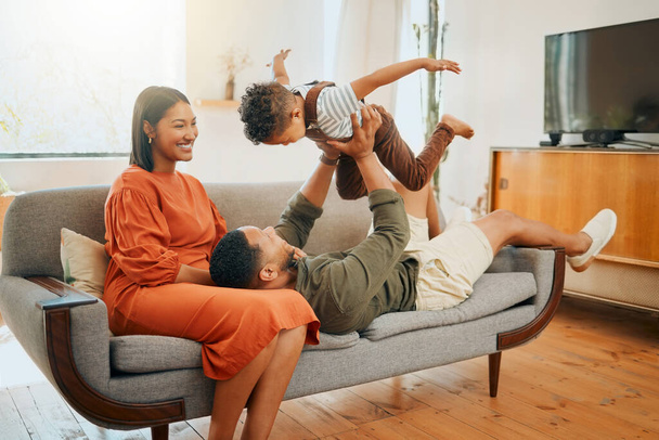 ラウンジでリラックスして一緒に遊んでいる3人の幸せな混合レース家族。自宅でソファの上で楽しいゲームをプレイしながら、彼らの息子と黒の家族の絆を愛する. - 写真・画像