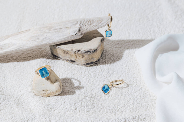 Elegant jewellery set of gold earrings and ring with blue topaz on white background with stones and wood. Minimal style composition. Product still life concept - Foto, immagini