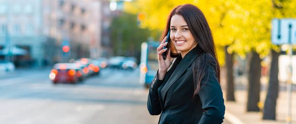 Mulher de negócios morena atraente em terno preto falando em um telefone celular em suas mãos ao ar livre.Copiar espaço.Banner, anúncio. - Foto, Imagem