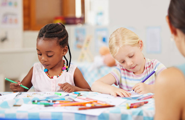 African american girl sitting at a table and colouring at pre-school or kindergarten with her caucasian classmate . Young female children using colourful pencils to draw pictures in a class at school. - Фото, изображение