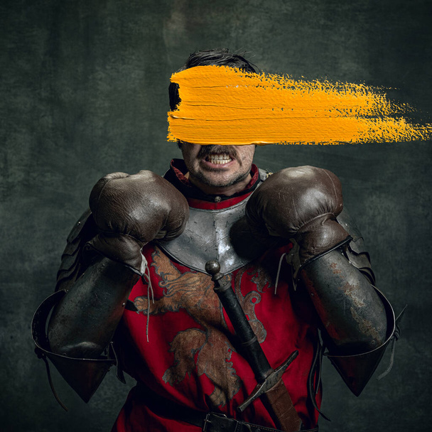 Power. Modern art design. Young man as medieval warrior, knight isolated on dark vintage background. Random stroke of yellow paint on his face. Emotions, comparison of eras, art, new vision concept. - Foto, imagen