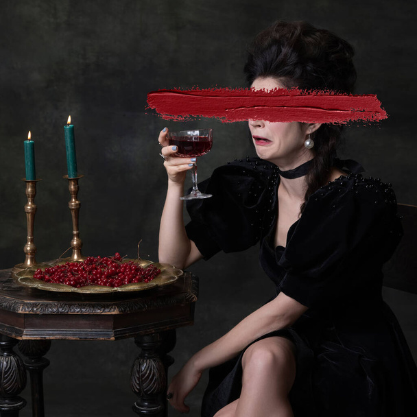 Tasting bitter wine. Hiding feelings. Young beautiful woman in image of medieval royal person in black dress isolated on dark vintage background. Concept of emotions, modernity and renaissance. - Photo, image