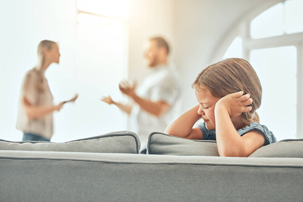 Scared little kid sitting and blocking her ears with hands while parents fight. Couple shouting at each other and arguing in presence of their daughter. Parent divorce affecting child mental health. - 写真・画像
