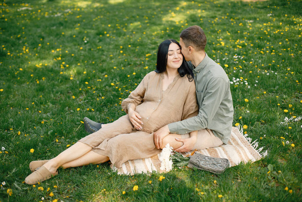 Couple waiting for their first baby sitting on a grass and hugging. Pregnant woman and her husband sitting in park and posing for a photo. Brunette woman wearing beige dress. - Foto, Imagem