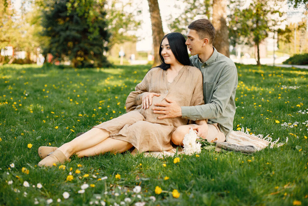 Couple waiting for their first baby sitting on a grass and hugging. Pregnant woman and her husband sitting in park and posing for a photo. Brunette woman wearing beige dress. - Fotoğraf, Görsel