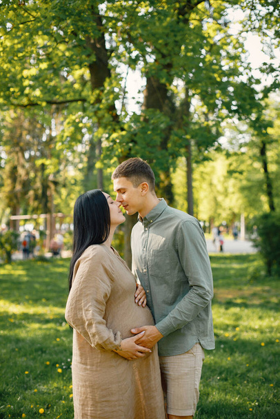 Couple waiting for their first baby standing on a grass and hugging. Pregnant woman and her husband standing in park and posing for a photo. Brunette woman wearing beige dress. - Φωτογραφία, εικόνα