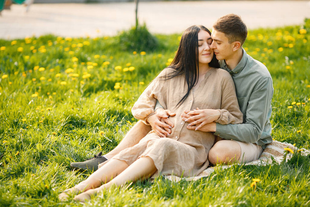 Пара ждет своего первого ребенка, сидящего на траве и обнимающегося. Беременная женщина и ее муж сидят в парке и позируют для фото. Брюнетка в бежевом платье. - Фото, изображение