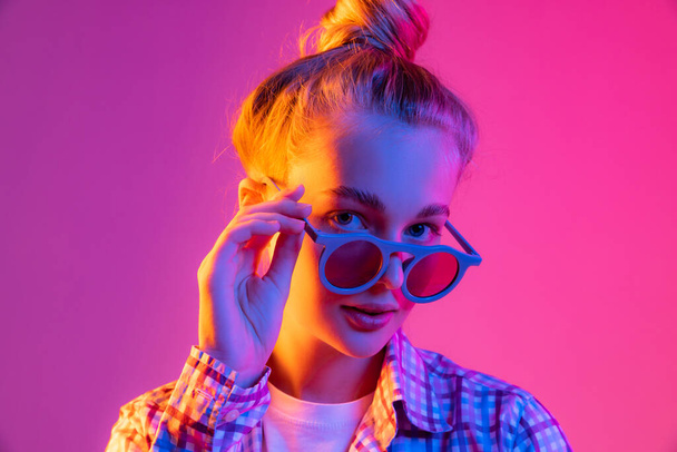 Party. Pretty young girl, student in cool eyeglasses wearing plaid shirt isolated on magenta color background in neon light. Concept of beauty, art, fashion, youth, sales and ad, education, studying. - Фото, изображение