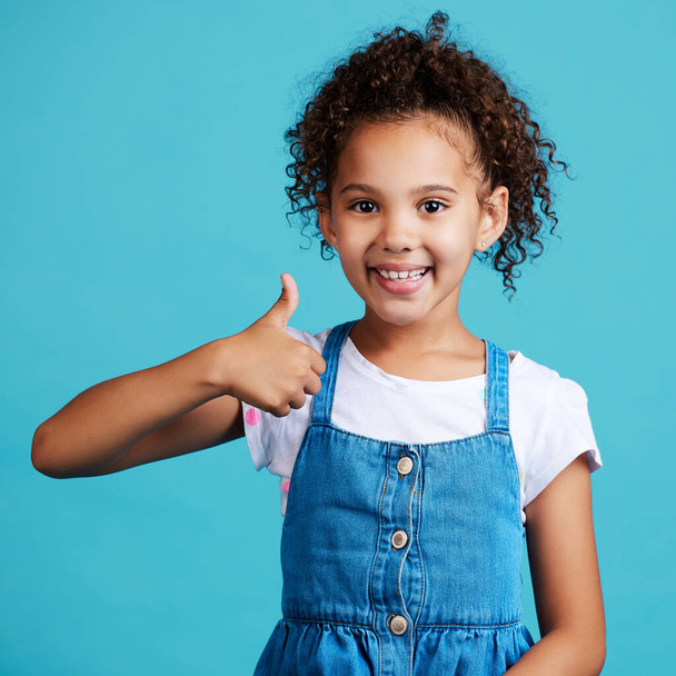 an adorable little girl showing thumbs up while standing against a blue background. - Foto, Imagen
