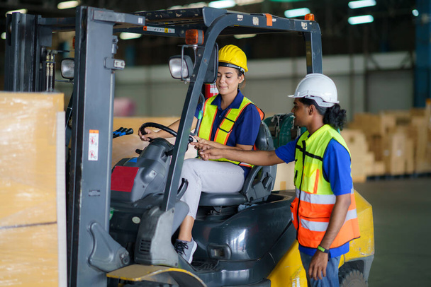 Asian man teaching woman driver drive fork lift car in industry with smile, ability of girl and diversity of career wearing helmet in reflective vest working on vehicle - Фото, изображение