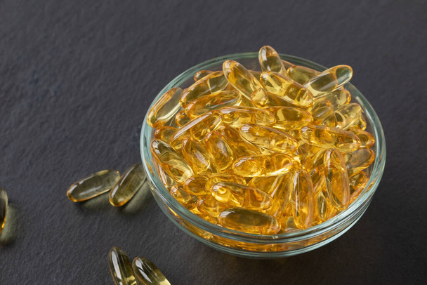 Cod liver fish oil capsules in glass bowl on black slate plate background. Omega 3, Vitamin E supplements. Healthy diet concept. Close-up, copy space - Foto, Imagen