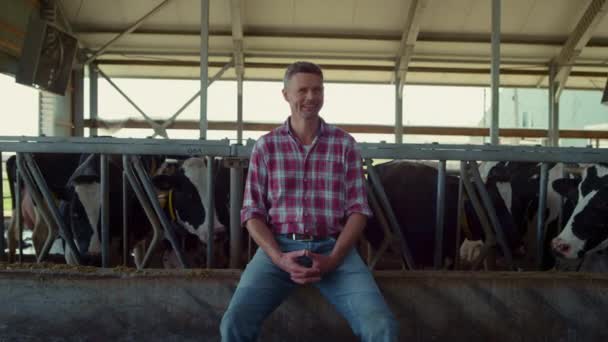 Happy farmer posing sitting on cowshed feeder near healthy cows. Charming professional agro worker enjoying work process in farmland barn. Middle age ranch owner smiling relaxing on modern farm. - Séquence, vidéo
