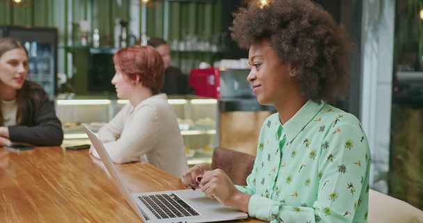 A happy black woman using laptop at coffee shop. One African American girl typing on computer keyboard smiling. A female entrepreneur person looking at laptop screen - Foto, Imagen