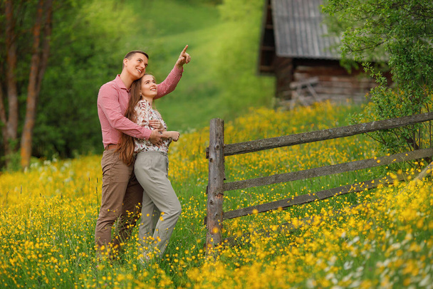Young loving couple gently hugging on the wood house background, a lot of yellow flowers are around on the field. Enjoying time together.  - Valokuva, kuva