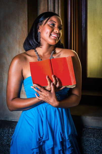 Lezen bij het raam. Jonge zwarte vrouw, met rood boek, bij het raam staan, lezen, glimlachen - Foto, afbeelding
