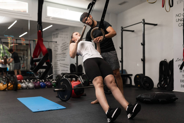 Young woman plays sports and performs single arm ring row and ring hold, at local training and fitness center, together with her personal trainer - Photo, image