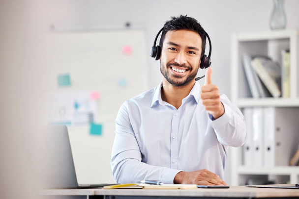 Young happy handsome mixed race male call center agent showing a thumbs up wearing a headset sitting at a desk in an office alone at work. Pleased hispanic customer service agent answering calls. - 写真・画像