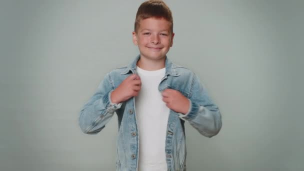Cheerful handsome happy toddler boy in jeans jacket smiling, looking at camera. Young fashion model teenager child kid schoolboy indoor studio shot isolated on gray background. Male nature beauty - Filmati, video