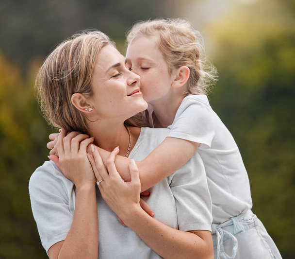 Szczęśliwa i zadowolona młoda matka przytulająca i obejmująca swoją córeczkę na zewnątrz w ogrodzie. Opiekuńcza dziewczynka stojąca całując matkę w policzek i przytulając ją w parku. - Zdjęcie, obraz
