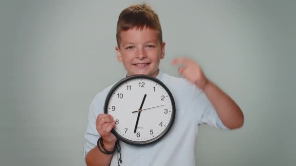 It is your time. Toddler kid boy showing time on wall office clock, ok, thumb up, approve, pointing finger at camera. Young teenager child schoolboy isolated alone on gray studio background indoors - Filmagem, Vídeo